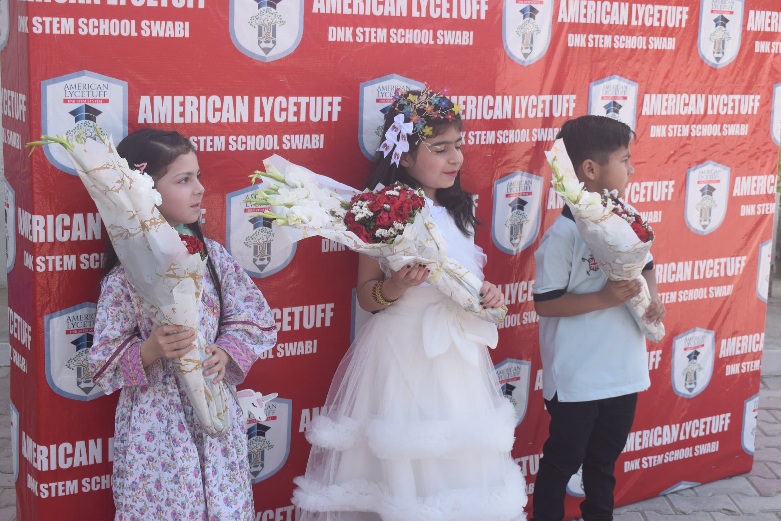 Young American Lycetuff Swabi students holding bouquets in front of the school banner